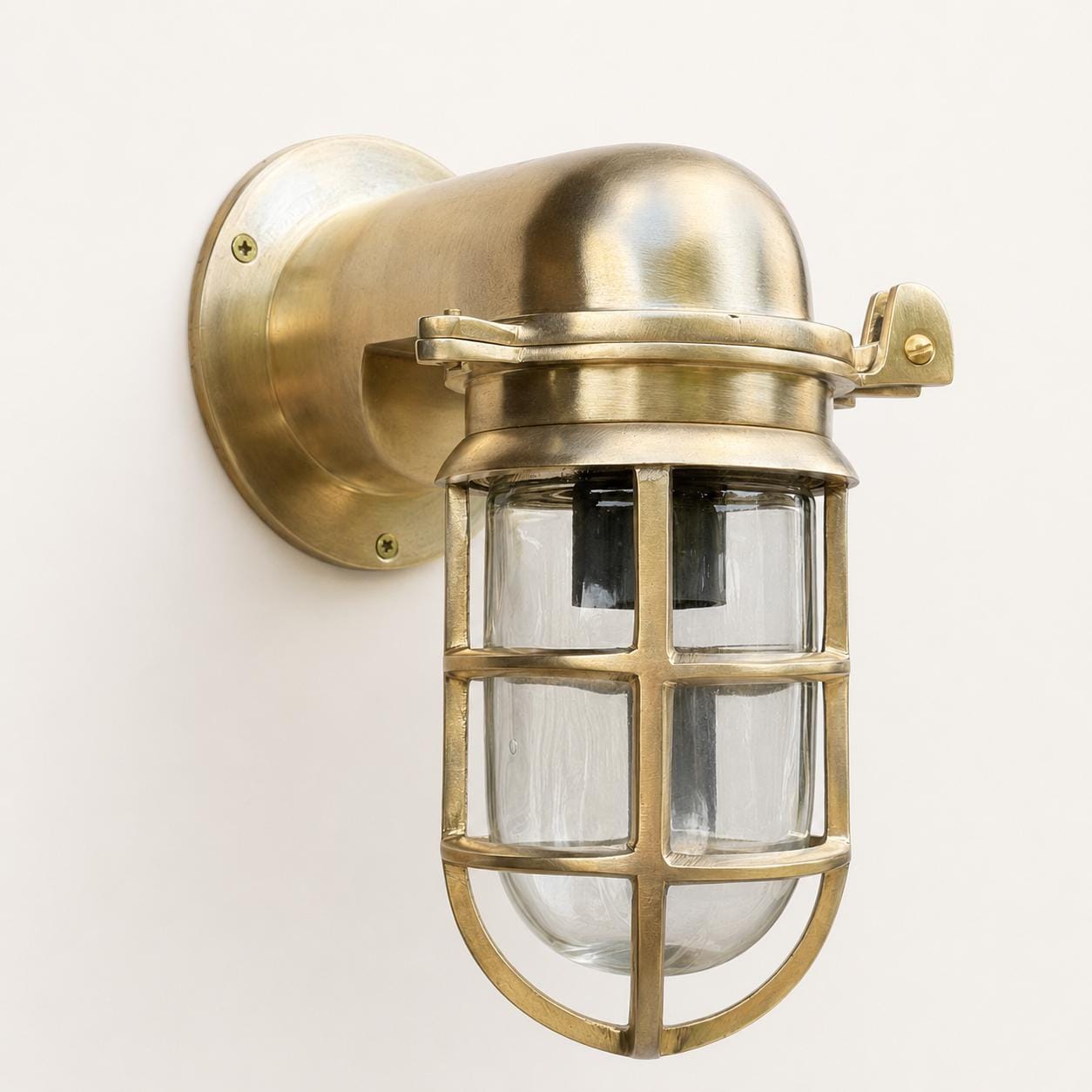 A classic industrial marine-style wall sconce with a brass-finished metal body, protective cage, and clear glass shade, mounted on a plain off-white wall.