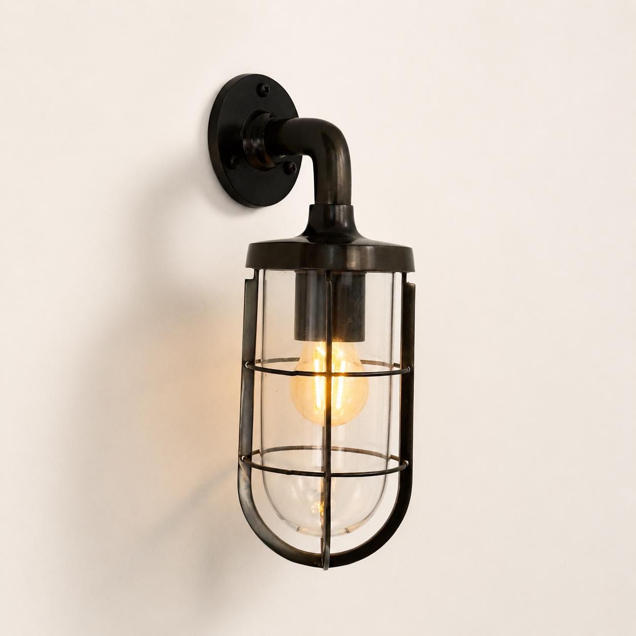 Industrial-style wall sconce with antique brass metal wall mount, curved arm, and clear glass cylindrical cage housing a glowing Edison bulb against a plain off-white wall.