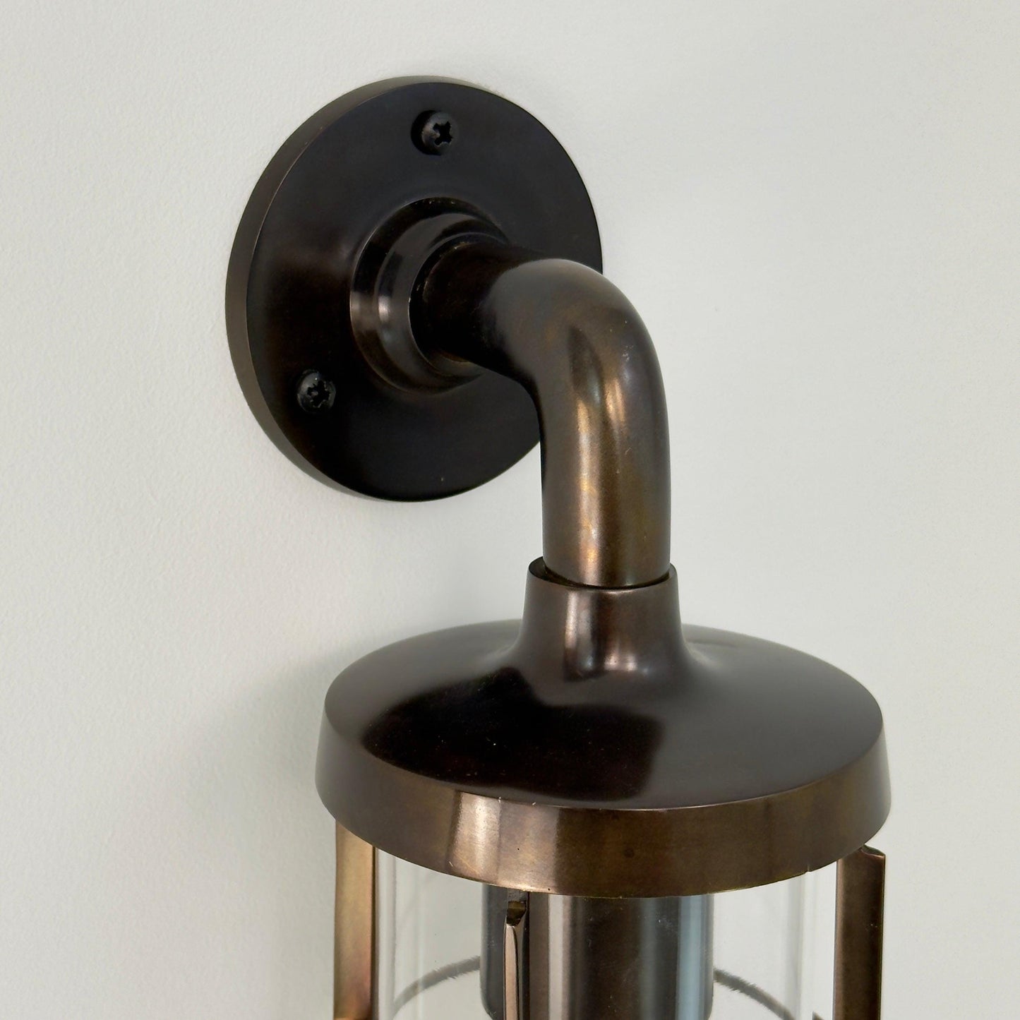 A close-up of a wall-mounted lantern light fixture with an antique genuine solid brass body, curved neck, round wall plate, and glass shade, shown against a plain off-white wall.